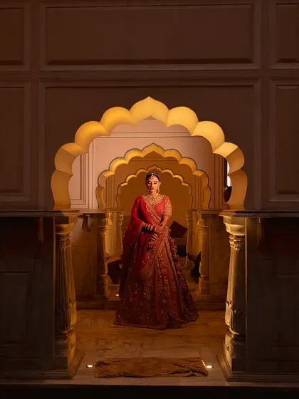 A bride stands framed by the magnificent scalloped arches of Alila Fort Bishangarh. The architectural beauty of the heritage venue, combined with soft lighting, creates a truly royal portrait.