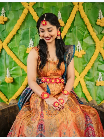 The bride, adorned with floral jewelry, smiles shyly during her Haldi ceremony against a backdrop of banana leaves.