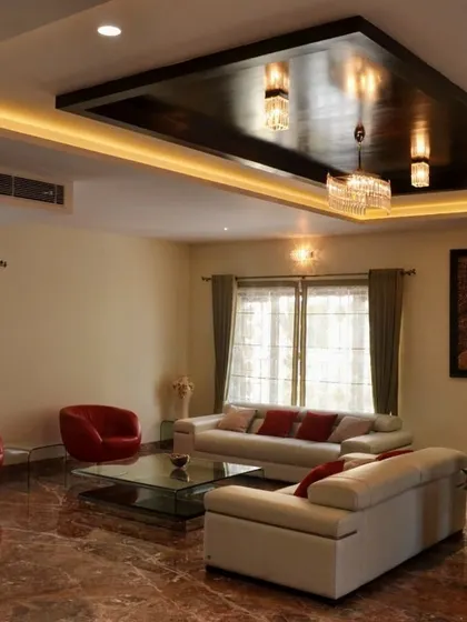 A modern living room featuring a striking black high-gloss false ceiling with integrated cove lighting and a chandelier. The space is furnished with minimalist white and red leather sofas, a glass coffee table, and set on a rich, red-toned marble floor.