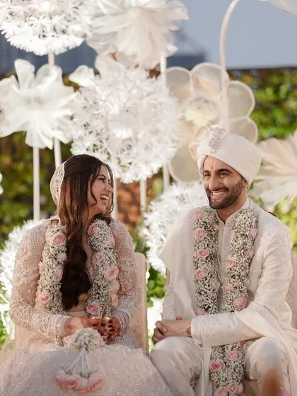A moment of shared laughter during the ceremony. The couple, dressed in elegant white, look at each other with genuine happiness, seated against a stunning floral backdrop.