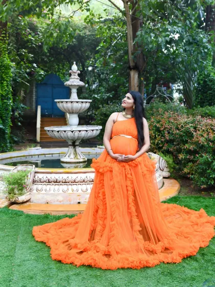 A grand portrait of a mom-to-be in an orange gown by a garden fountain, looking up peacefully.