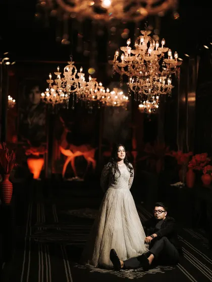 A wide-angle shot capturing the couple in a dramatic, low-lit setting. The magnificent chandeliers hanging above them add to the grandeur and moody atmosphere of their Sangeet.