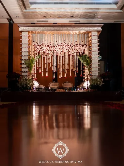 A traditional red and white mandap setup inside a grand hall. The decor features pillars adorned with diyas and a ceiling covered in hanging garlands of jasmine and red roses.