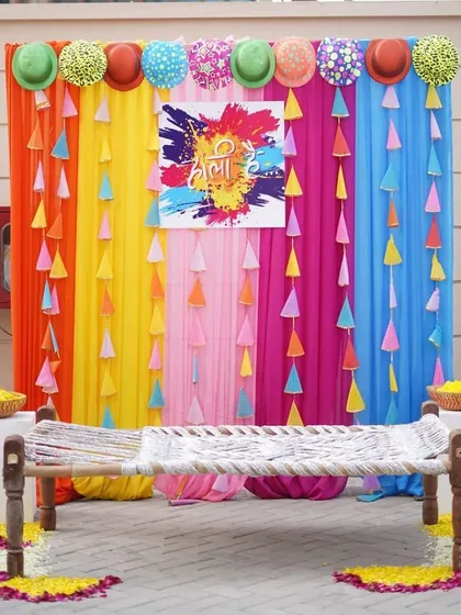 A colorful and rustic photo booth for a Holi party. The backdrop of rainbow curtains and a traditional charpai (woven cot) perfectly blends modern colors with village fun.