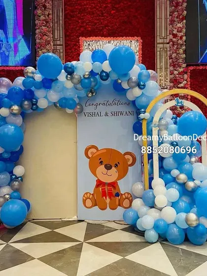 This large-scale baby shower decoration was set up in a banquet hall. It features a double balloon arch in shades of blue and silver, with a custom backdrop featuring a cute teddy bear and the parents' names.