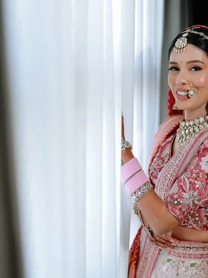A beautiful bridal portrait by the window. The natural light highlights the bride's features and the delicate embroidery on her pink and red lehenga.