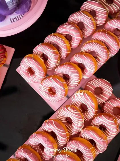 A delicious-looking spread of pink-frosted donuts with white chocolate drizzle, perfectly aligned on serving trays. These treats are always a hit and add a pop of color to any dessert table.