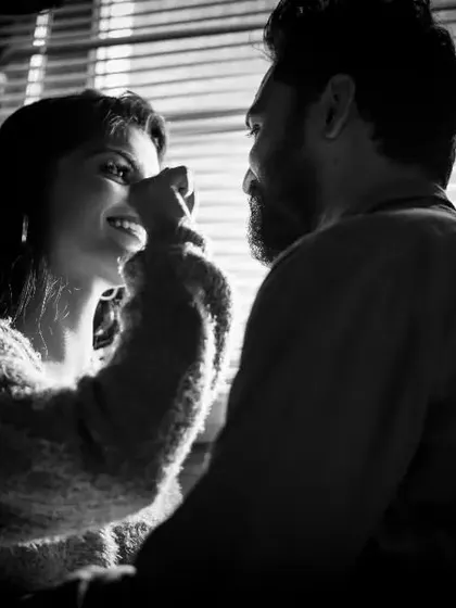 A playful and loving moment between a couple, captured in beautiful black and white. The light from the window blinds creates a lovely pattern, adding texture and mood to this candid shot.