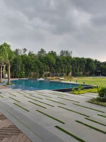 The expansive swimming pool of a Delhi farmhouse, featuring a modern design with a wooden deck and stone pavers with grass joints. The view extends across a vast lawn, creating a sense of openness and luxury.