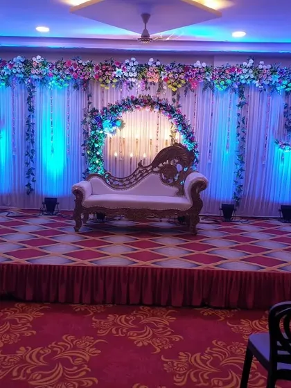 A classic engagement stage decoration in a banquet hall. The backdrop features white drapes with a floral border, enhanced by blue uplighting and decorative white trees.