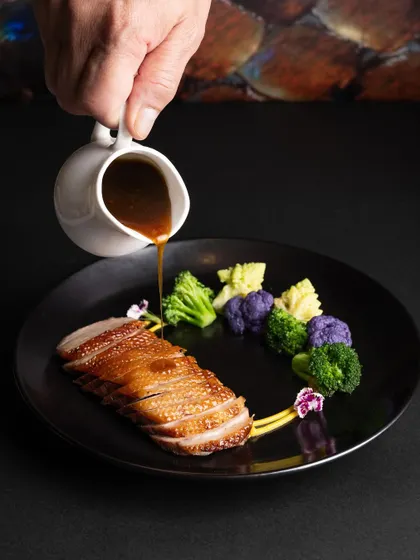 A fine-dining plate of sliced duck breast with a sauce being poured, capturing a moment of culinary action and elegance.