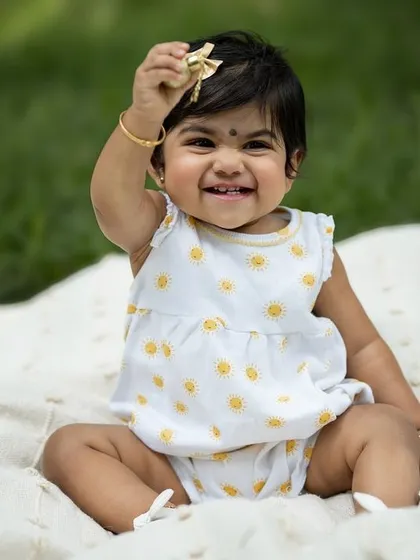 A baby girl sits on a blanket outdoors, smiling brightly and holding up a small toy. A simple, happy moment captured in the sunshine.
