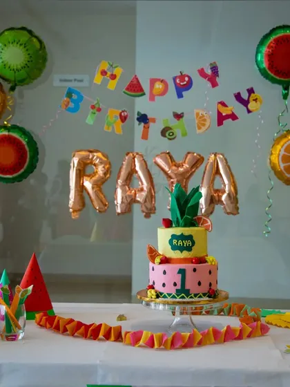 The complete cake table for Raya's fruit-themed first birthday. The setup includes a custom cake, fruit-shaped mylar balloons, and a colorful "Happy Birthday" banner.