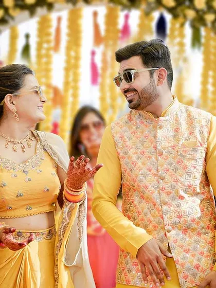 A fun, candid shot from a haldi ceremony. The couple, wearing sunglasses and bright yellow outfits, share a laugh, capturing the playful and joyous spirit of the event.