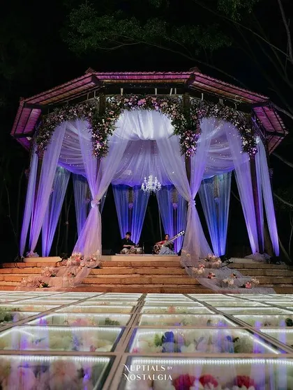 A breathtaking Sangeet stage featuring a glass floor filled with flowers. The mandap structure is draped in ethereal white and purple fabrics, lit to create a truly magical and one-of-a-kind dance and performance area.