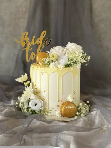 An elegant "Bride to Be" cake with a white chocolate drip, fresh white flowers, and gold macarons. This design is sophisticated and perfect for a bridal shower.