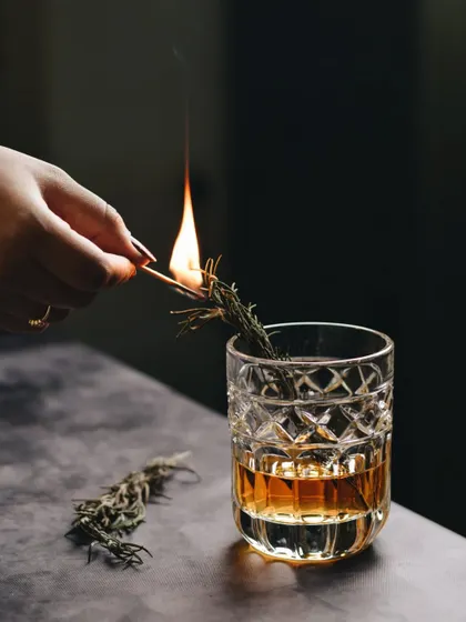 The moment of ignition. This shot captures the drama of lighting a rosemary sprig for a smoked cocktail, showcasing a technique that adds aroma and flair.