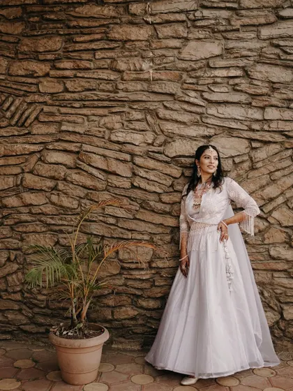 A full-length bridal portrait against a rustic stone wall. The bride's elegant white gown and confident pose create a stunning and timeless image.