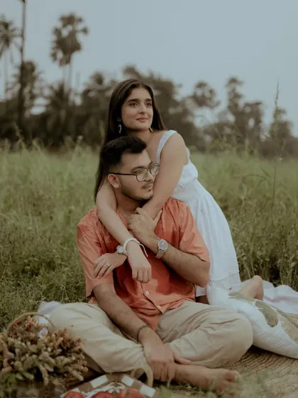 A classic romantic pose, with her embracing him from behind as they sit together in the field.