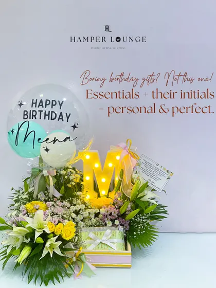 Personalization makes any gift feel more special. This birthday hamper for Meena features her initial 'M' in a bright marquee light, surrounded by a lush arrangement of yellow and white flowers and a custom "Happy Birthday Meena" balloon.