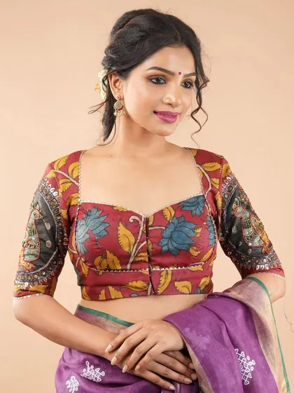 This sweetheart neck blouse features a beautiful floral Kalamkari design on a deep red base. The sleeves have a contrasting black panel with their own unique motif, adding to its artistic appeal.