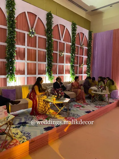 A beautiful and functional seating area for a Mehendi. The custom backdrop features a pastel color-blocked wall with elegant window-pane frames and columns of greenery, set against a vibrant floral carpet.