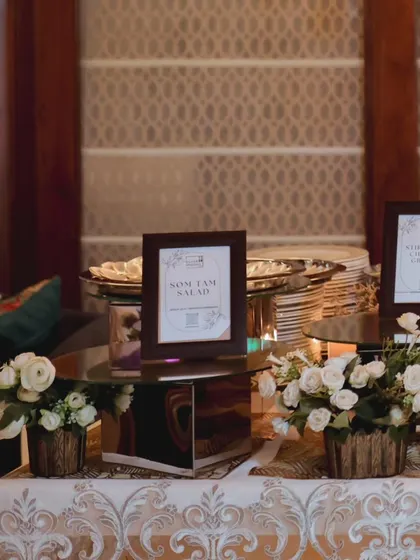An elegant buffet setup for a birthday celebration, featuring mirrored stands, white floral arrangements, and framed menu cards for a clean and sophisticated look.