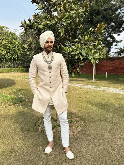 A full-length view of the Sikh groom's ensemble. The ivory sherwani, crisp white churidar, and traditional juttis come together for a look that is both timeless and impeccably stylish.