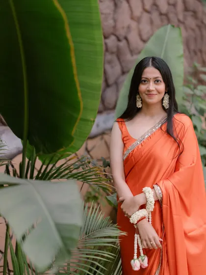 A graceful portrait of the bride in her orange saree, adorned with floral jewelry, posing against a backdrop of lush green leaves.