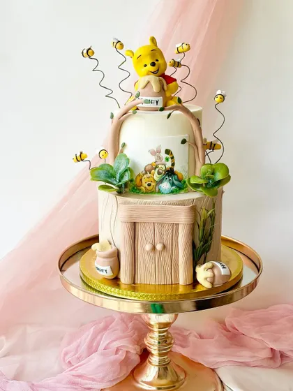 A detailed Winnie the Pooh cake set in the Hundred Acre Wood. This two-tiered creation features Pooh Bear with his honey pot, Piglet, and buzzing bees, with a charming wood-paneled door and fondant leaves.