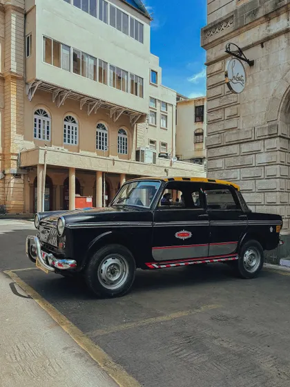 A classic black-and-yellow taxi parked in a quiet spot. The composition highlights the car's distinct shape and its role as a Mumbai icon.