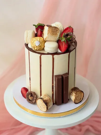 A fully loaded drip cake for the ultimate treat lover. This cake features a chocolate drip and is topped with toasted marshmallows, strawberries, Ferrero Rocher, KitKats, and macarons.