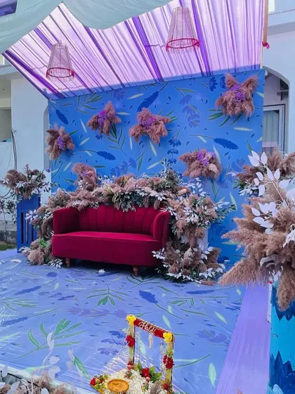 A unique Haldi stage with a blue and lavender color theme. The backdrop has a floral print, and the stage is decorated with dried pampas grass for a rustic touch.