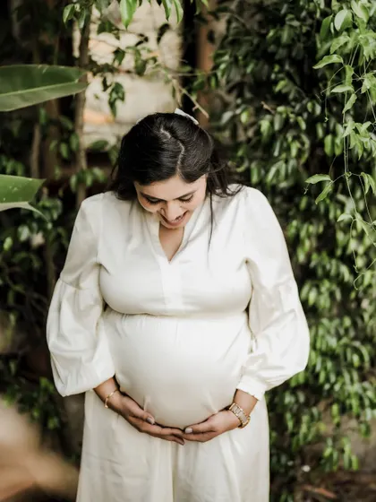 A happy and radiant portrait of the mom-to-be cradling her bump, surrounded by lush green leaves. Her smile is full of joy.