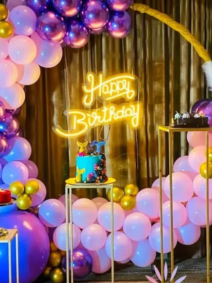 A chic birthday setup with a circular balloon arch in shades of purple, pink, and gold. The "Happy Birthday" neon sign adds a modern, celebratory glow to the party.
