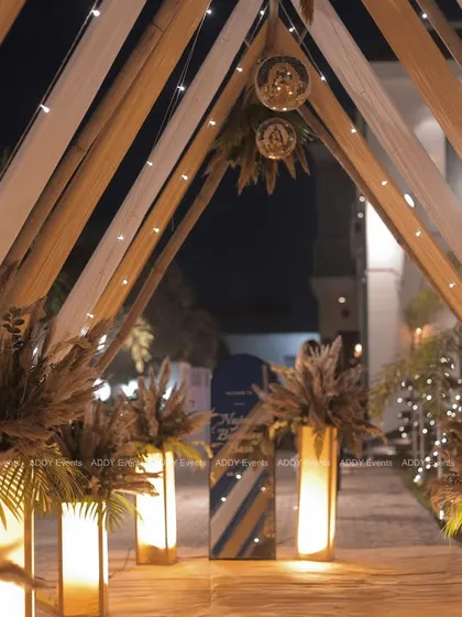 For an evening birthday bash, we created this stunning boho-glam entrance walkway. The triangular arch, draped in fabric and lit with string lights, is lined with illuminated pillars topped with pampas grass, with disco balls adding a touch of party sparkle.