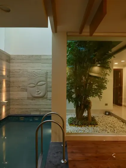 The stilt floor pool area at Gupta Residence is a tranquil retreat. A stone-clad wall with soft lighting and a Buddha sculpture is complemented by a glass-enclosed courtyard, creating a serene interplay of water, light, and greenery.