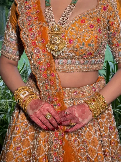 A detail shot focusing on the intricate embroidery of the lehenga and the bride's henna. The overall color palette is warm, bold, and beautifully traditional.