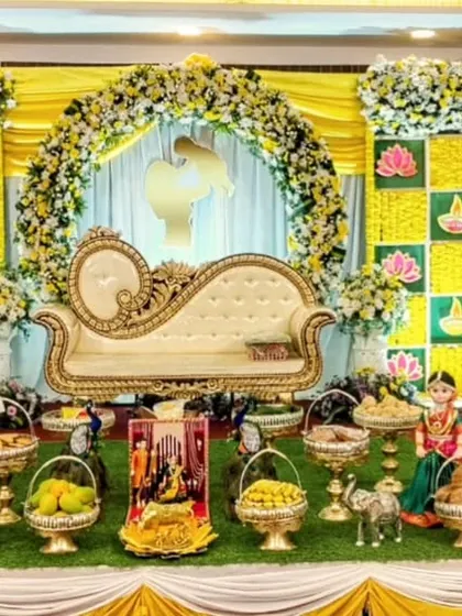 A vibrant yellow-themed baby shower stage, complete with a floral arch, traditional checkered backdrop, and a full display of fruits and sweets for the ceremony.