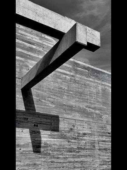 A detail of a sculptural concrete beam cantilevering from a board-formed wall. This black and white photograph emphasizes the form and the stark contrast of shadow against the textured surface.