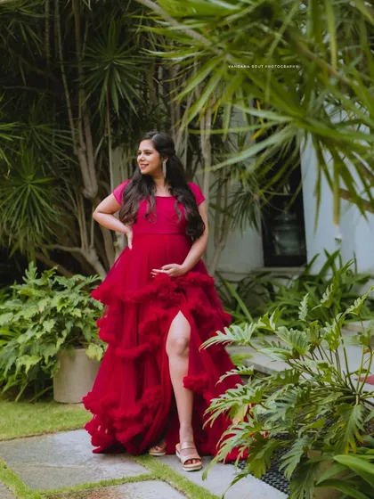 A full-length shot of a mother-to-be in a striking red gown with a thigh-high slit, posing elegantly amidst lush green plants.