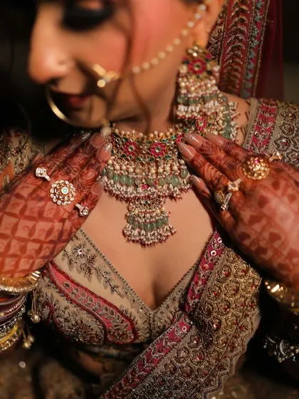 A close-up on the details. This image highlights a bride's henna-adorned hands as she adjusts her magnificent layered green and ruby Kundan necklace.