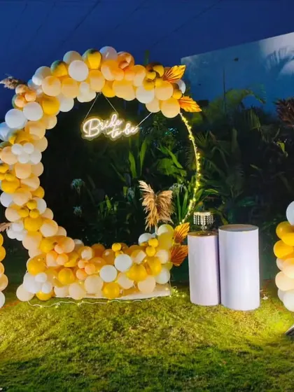 We take the party outdoors with creative setups like this 'Bride to be' celebration. A circular balloon arch in sunset yellows and whites, accented with a neon sign and prop champagne bottle, makes for a perfect garden party backdrop.
