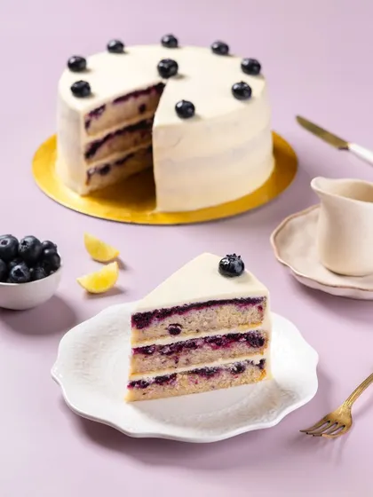A clean, bright shot of a layered blueberry lemon cake for The Big Chill Cafe. This image is styled for catalog and social media use, emphasizing the cake's structure and fresh ingredients against a simple, colorful background.