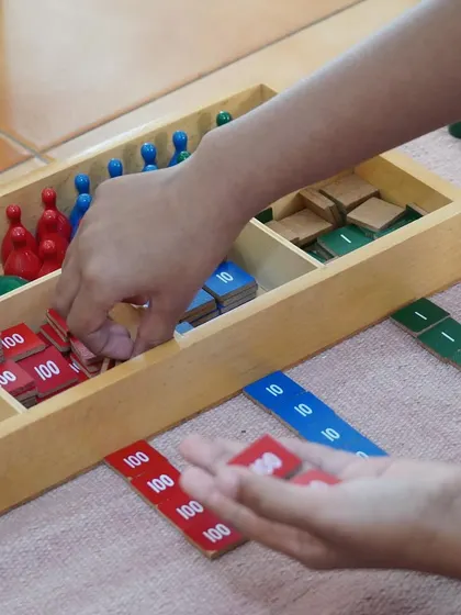A child's hands sort through stamp game tiles, a material used for understanding the four mathematical operations. This shows how we introduce complex arithmetic in a simple, hands-on way.