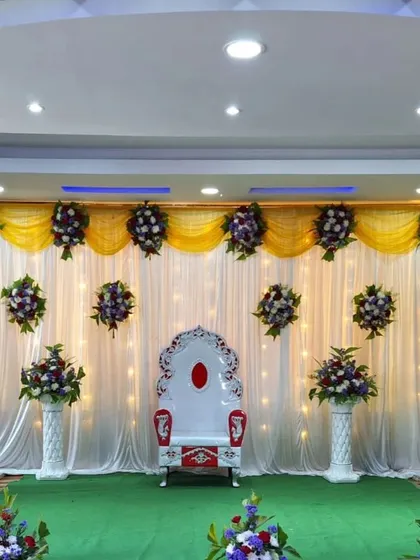 A simple yet regal setup for a traditional ceremony. The stage features a single ornate throne against a backdrop of white and yellow drapes with scattered floral bouquets, ensuring the focus remains on the main event.