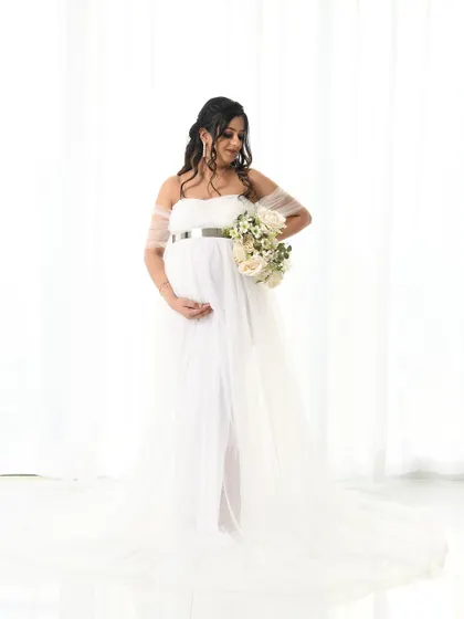 An elegant solo portrait of the mom-to-be in a white off-shoulder gown, holding a bouquet of white roses. The clean, high-key background emphasizes her graceful silhouette.