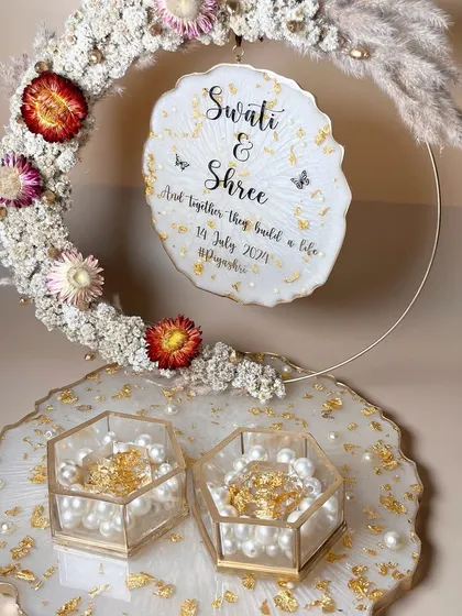 An elegant white and gold ring platter with a floral hoop featuring dried flowers and pampas grass. The hexagonal ring boxes are filled with pearls and gold flakes.
