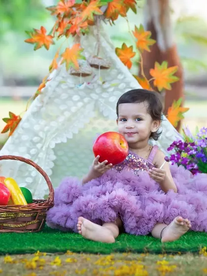 A sweet toddler in a fluffy purple dress sits in front of a teepee, holding an apple with a curious smile. The colorful props and natural setting make for a magical portrait.