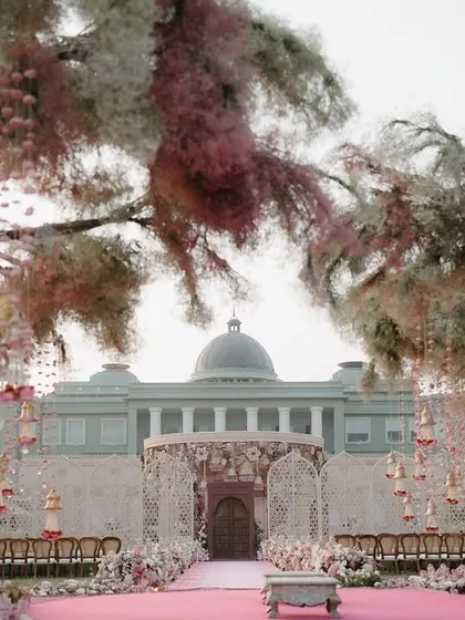 A grand mandap at Raffles Udaipur, with trees adorned in pink and white florals. The design creates a fairytale-like atmosphere, framing the palace in the background.